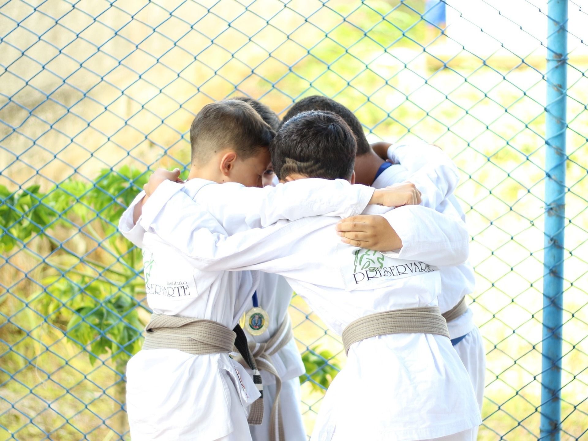 Imagem de 4 crianças aparentandoi ter 6 a 8 anos, abraçadas em círculo vestindo os quimonos brancos de jiu-jitsu do instituto preservarte em clima de concentração em um campeonato de Jiu-jitsu.